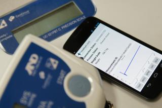 Ein Smartphone zeigt medizinische Daten neben einer digitalen Waage und einem medizinischen Gerät an, was auf eine Fernüberwachung des Gesundheitszustands oder den Einsatz von Telemedizintechnologie schließen lässt.