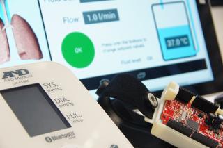Nahaufnahme von medizinischen Geräten, darunter ein Blutdruckmessgerät und ein elektronischer Schaltkreis, mit einem Computerbildschirm im Hintergrund, auf dem Lungengrafiken und Gesundheitsdaten wie Durchflussrate und Temperatur angezeigt werden.