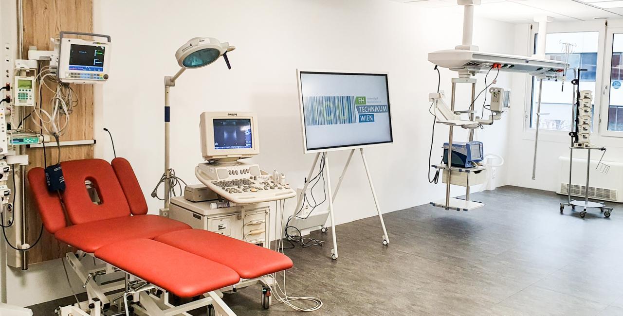 Ein modernes medizinisches Untersuchungszimmer mit einem roten Patientenstuhl, medizinischen Geräten, darunter Monitore und ein Ultraschallgerät, und einem großen Bildschirm, auf dem die Technische Woche angezeigt wird. Der Raum hat weiße Wände und große Fenster.