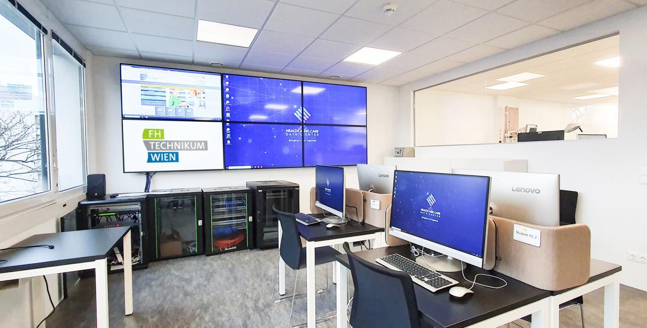 Ein helles, modernes Computerlabor mit mehreren Desktop-Computern auf Schreibtischen, großen, an der Wand befestigten Monitoren, die digitale Inhalte anzeigen, und Glasfenstern, die den Blick auf einen angrenzenden Büroraum freigeben. Auf den Schildern steht FH Technikum Wien.