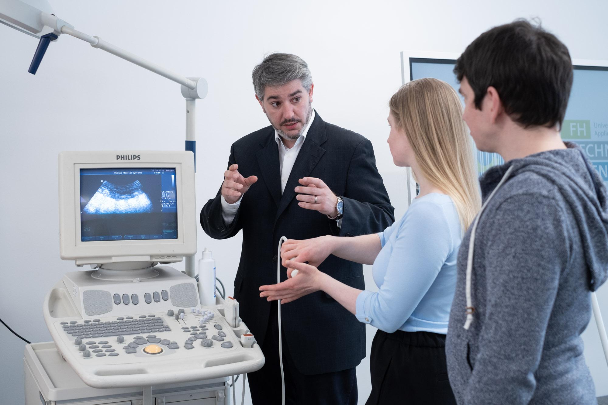 Ein Mann im Anzug erklärt einer jungen Frau, die den Schallkopf hält, die Bedienung eines Ultraschallgeräts, während ein junger Mann zuschaut. Auf dem Bildschirm wird ein Ultraschallbild angezeigt. Alle drei befinden sich in einer klinischen Umgebung.