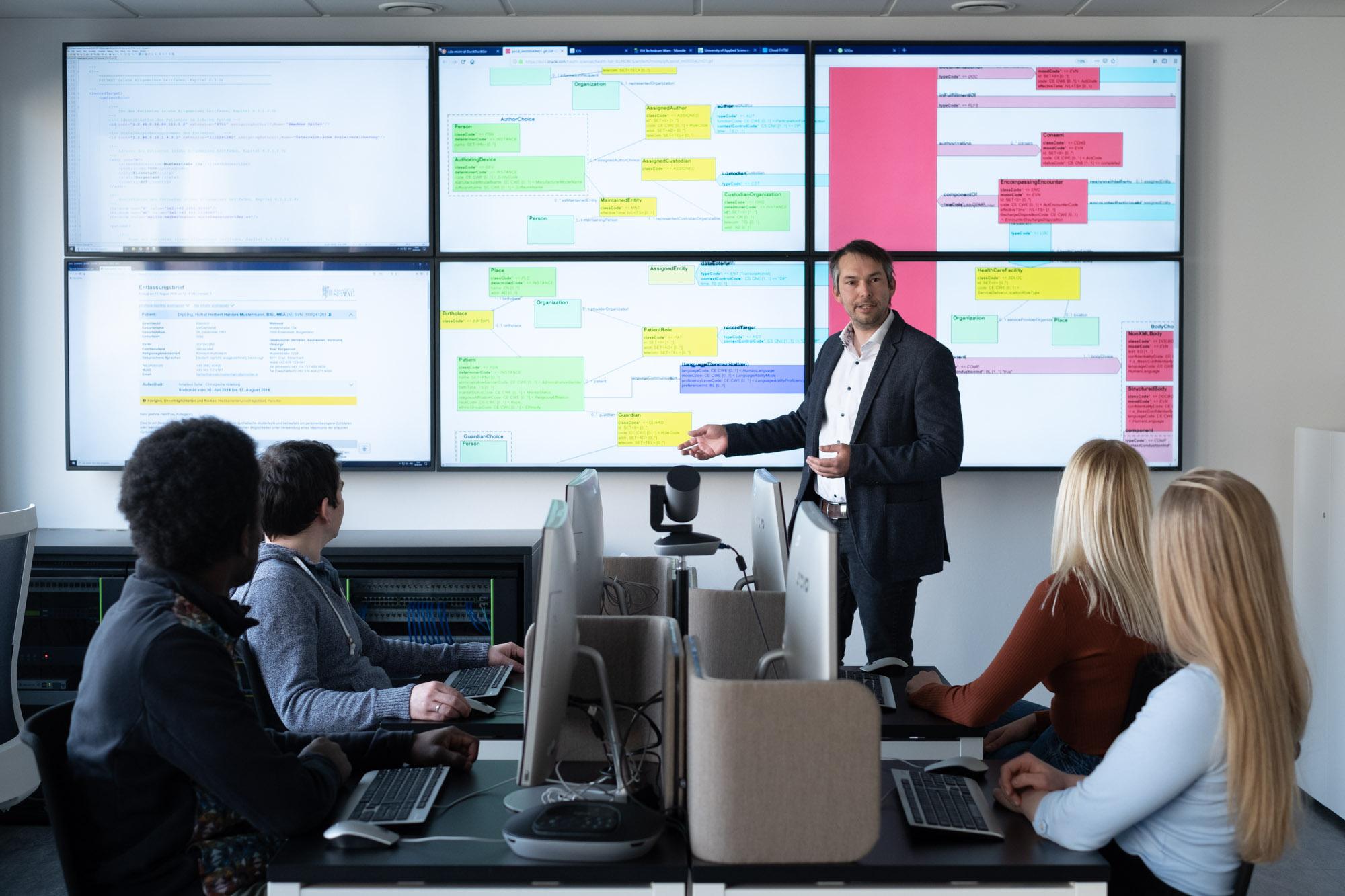 Ein Mann im Anzug präsentiert Flussdiagramme und Daten auf großen Bildschirmen vor vier Personen, die in einem modernen Büro oder Klassenzimmer an Computern sitzen.