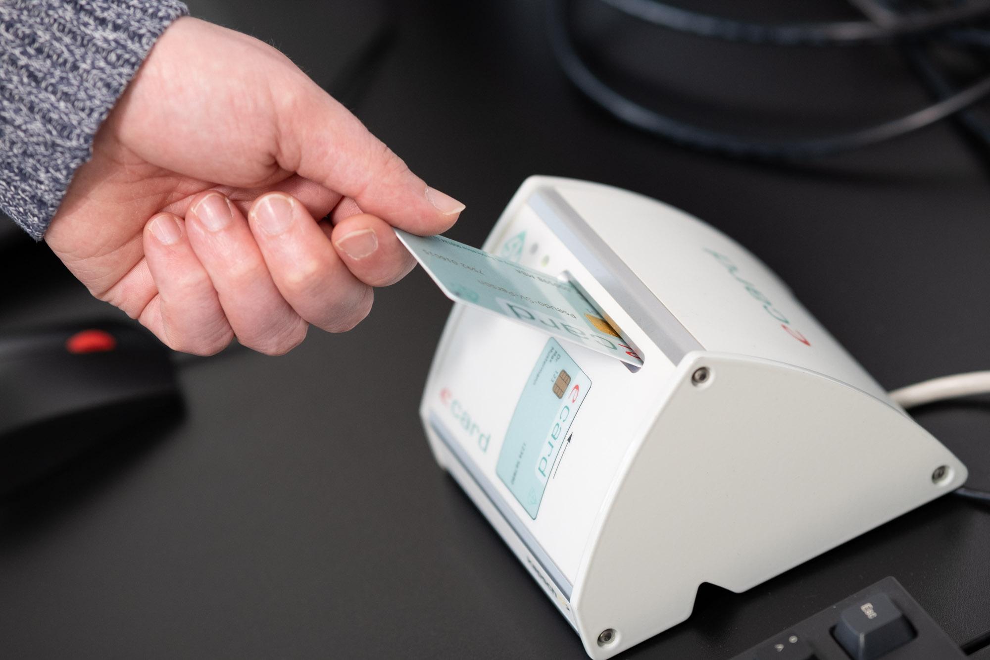 Eine Person steckt eine Ausweiskarte in ein weißes Kartenlesegerät auf einem schwarzen Schreibtisch; im Hintergrund sind Computerkabel zu sehen.