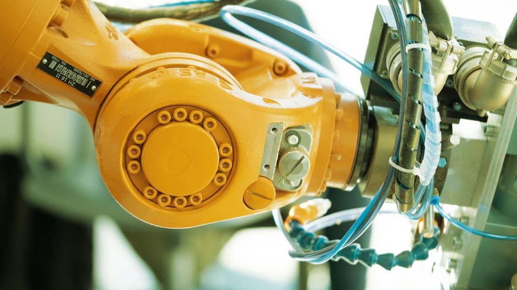 Nahaufnahme eines Industrieroboterarms mit orangefarbenen Gelenken und verschiedenen Kabeln, der in einer hell erleuchteten Umgebung Maschinen bedient.