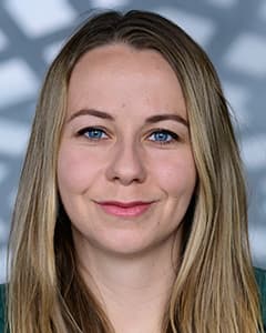 Eine Frau mit langen, glatten blonden Haaren und blauen Augen lächelt leicht. Sie trägt ein dunkles Oberteil und ist vor einem Hintergrund mit einem geometrischen, unscharfen Muster fotografiert.