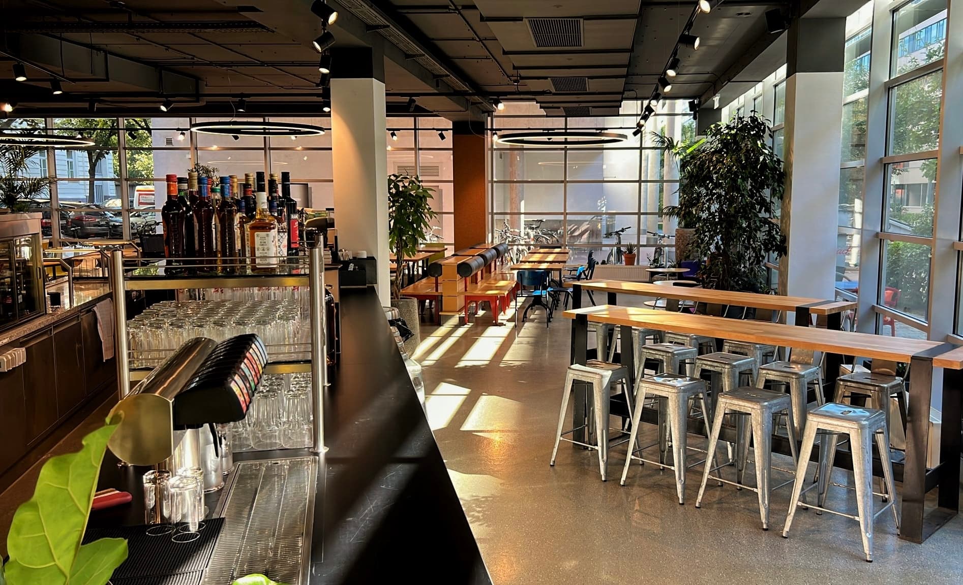 Eine moderne Bar mit hohen Tischen und Metallhockern, Flaschen auf dem Tresen und großen Fenstern, die das Sonnenlicht hereinlassen. Grünpflanzen schmücken den Raum, und leere Tische warten auf Kunden.