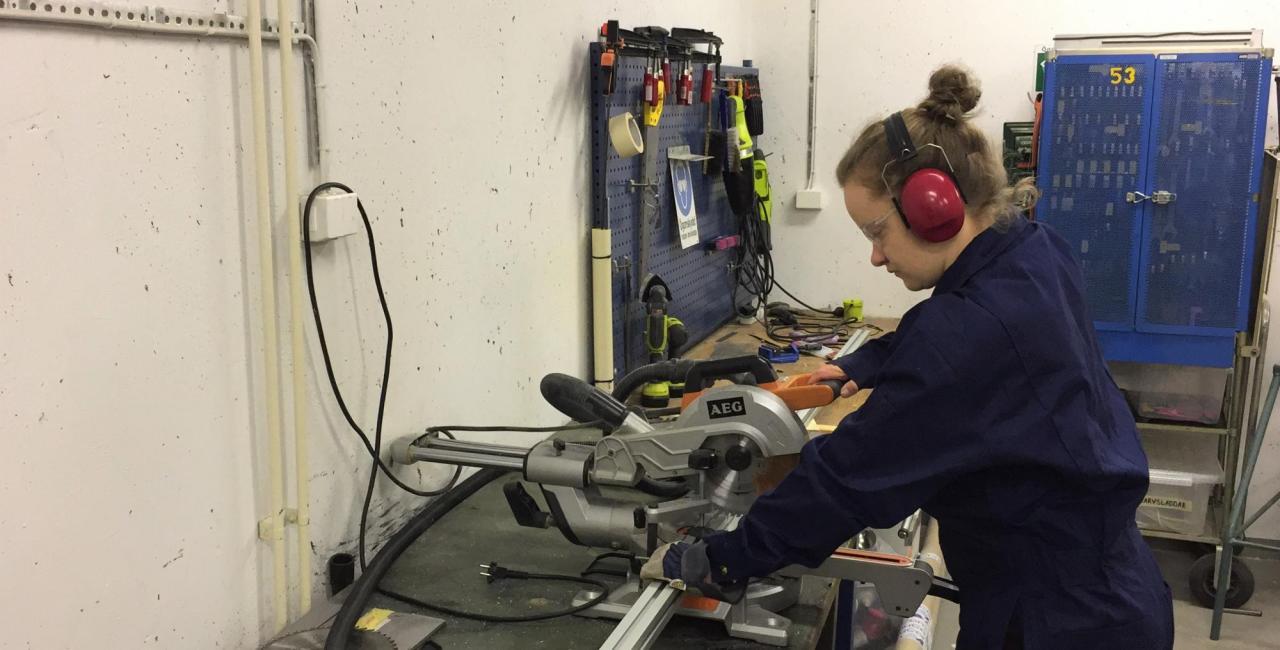 A person wearing ear protection and a navy blue coverall operates a circular saw at a workbench in a workshop with tools hanging on the wall behind them.