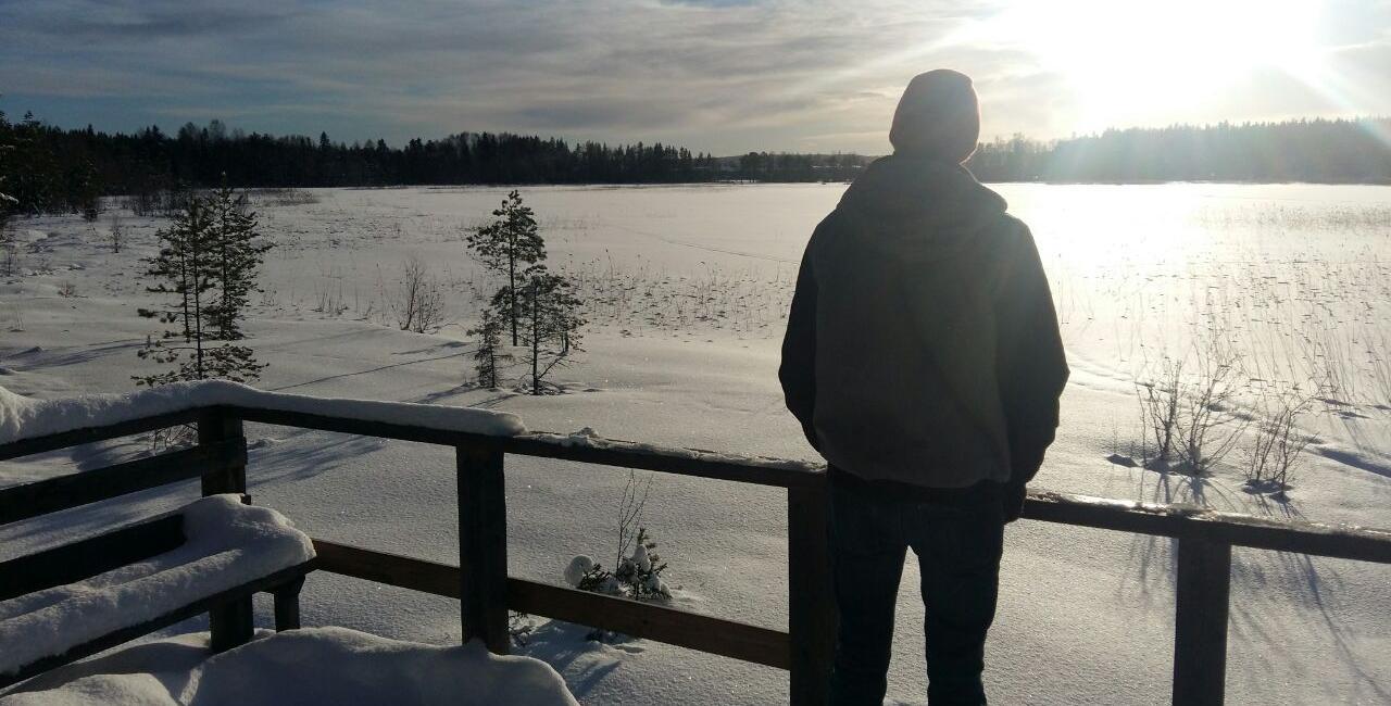 A person in a hoodie stands on a snowy wooden deck, looking out over a frozen, snow-covered landscape with sparse trees and bright sunlight low in the sky.