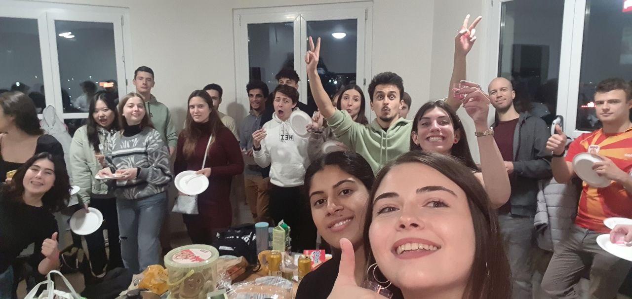 Eine Gruppe junger Erwachsener lächelt und posiert für ein Selfie auf einer Party. Einige halten Teller und Tassen, und auf dem Tisch vor ihnen steht Essen. Die Umgebung scheint ein Innenraum mit großen Fenstern im Hintergrund zu sein.
