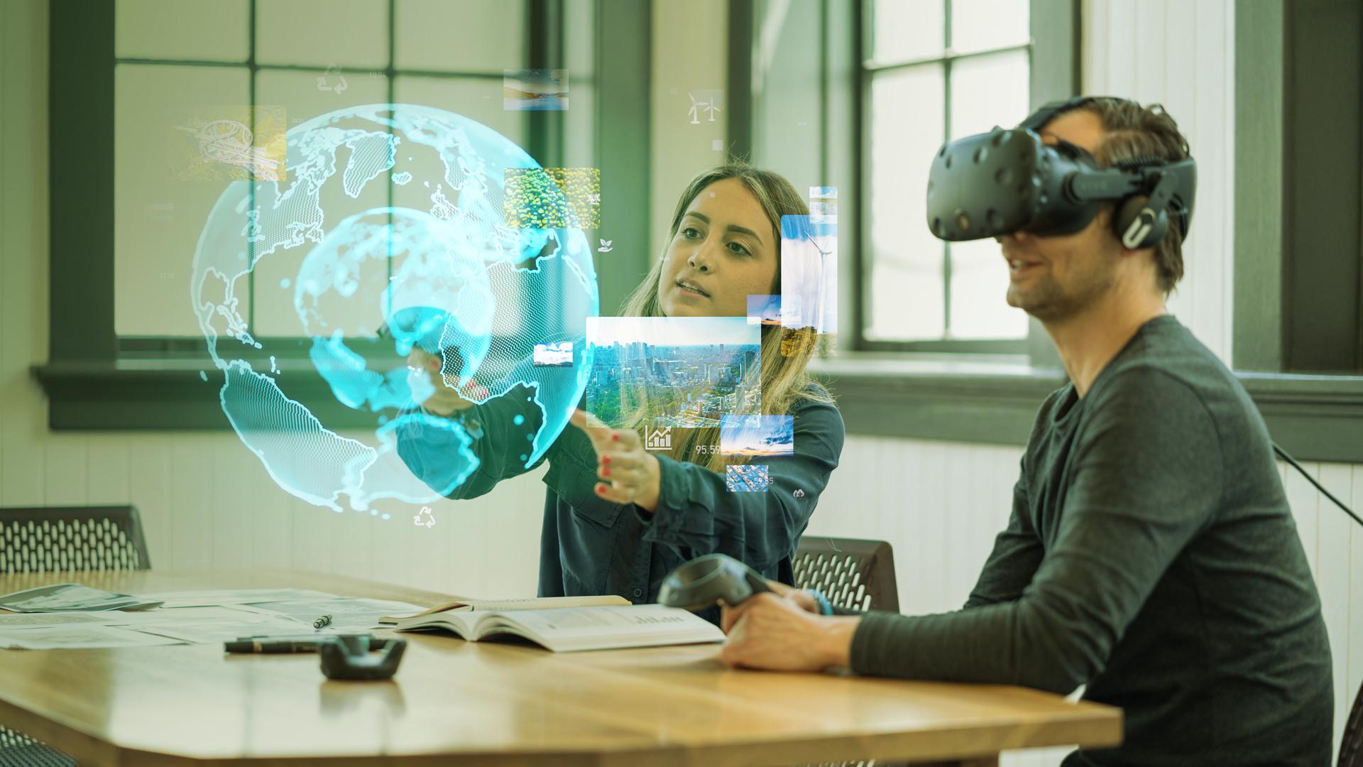 Zwei Personen sitzen an einem Tisch und tragen Virtual-Reality-Headsets. Ein digitaler 3D-Globus und verschiedene Bilder werden in die Luft projiziert, was darauf hindeutet, dass sie mit Augmented-Reality-Technologie interagieren.