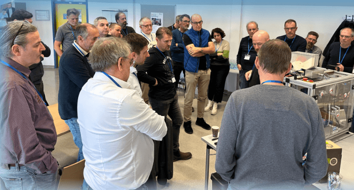 Eine Gruppe von Menschen steht um einen Präsentationstisch herum und verfolgt aufmerksam, wie eine Person in einem hellen Raum ein wissenschaftliches oder technisches Gerät erklärt.